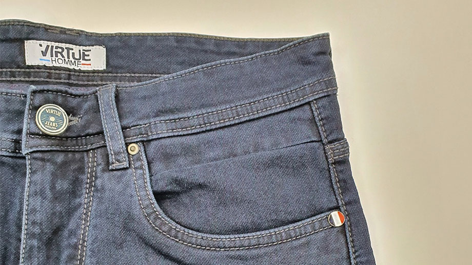 Photo d'un jean de la marque Virtue. Le jean n'est pas porté et est d'un bleu tendant vers le gris le fond derrière le jean est jaune pâle.
